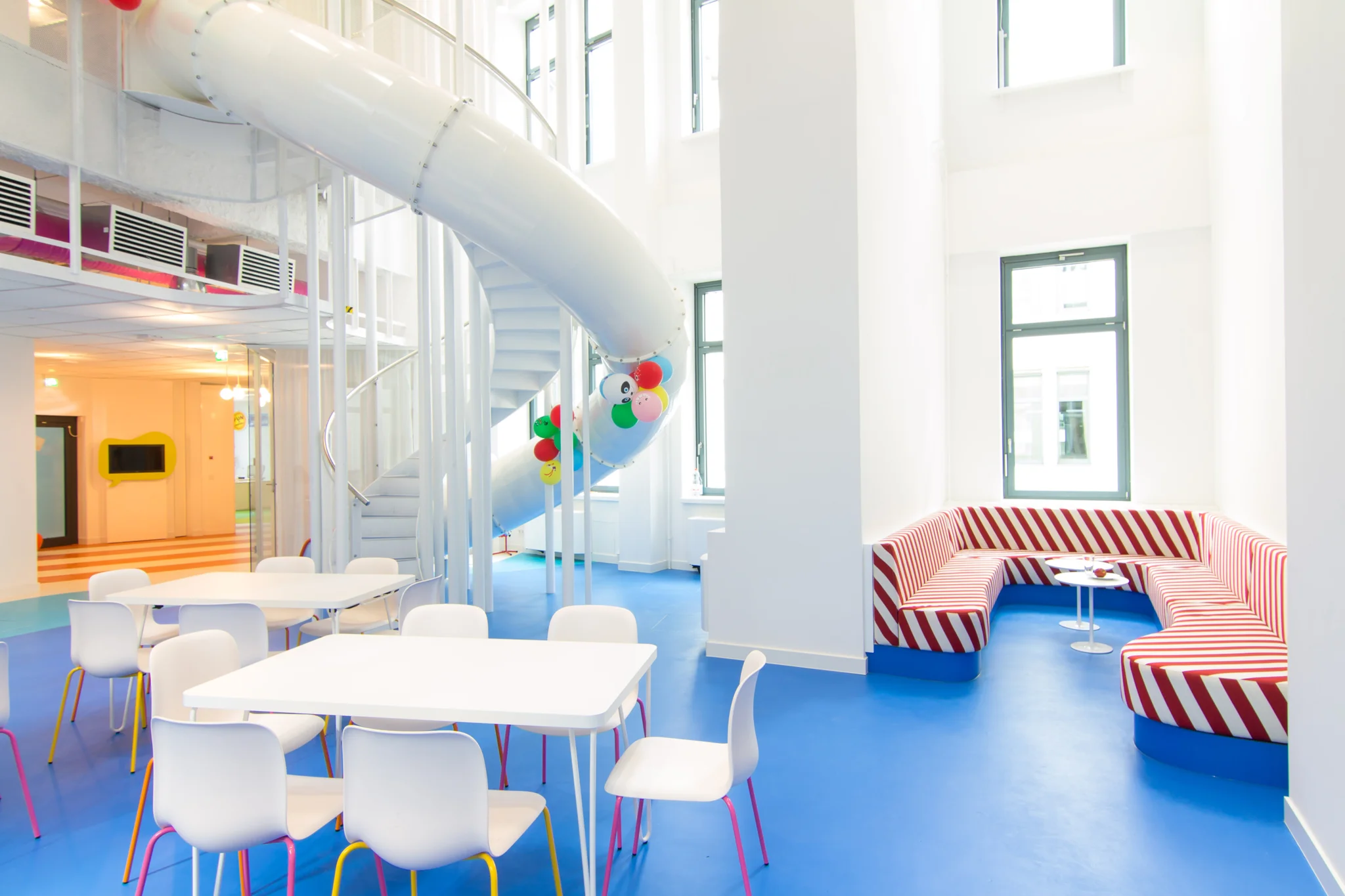 blue flooring and white double-height space in a cafe area with a white slide, red and white striped sofa