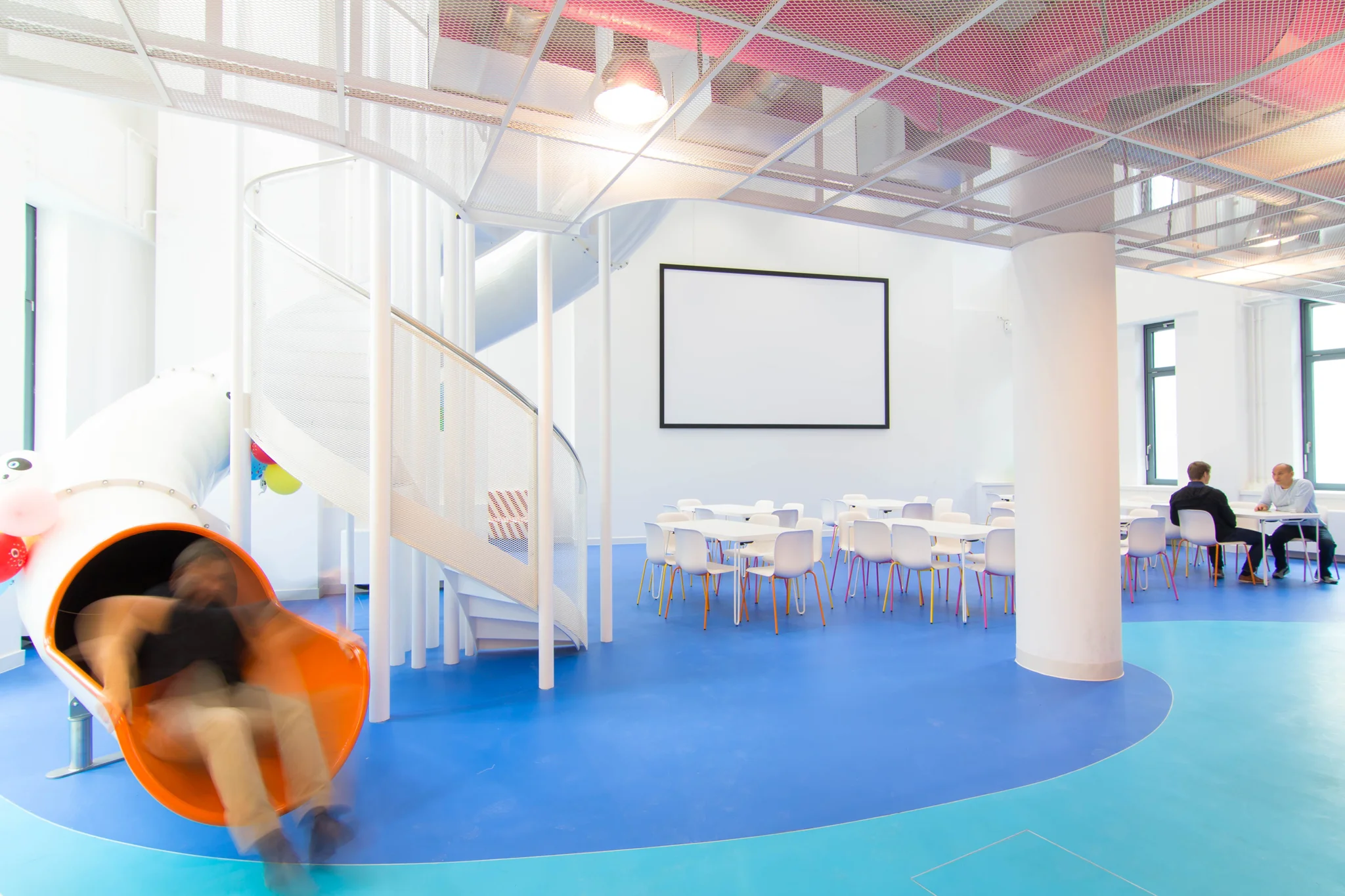 A man exiting a white slide/flume onto a blue flooring area and white double-height space in a cafe area with white furniture