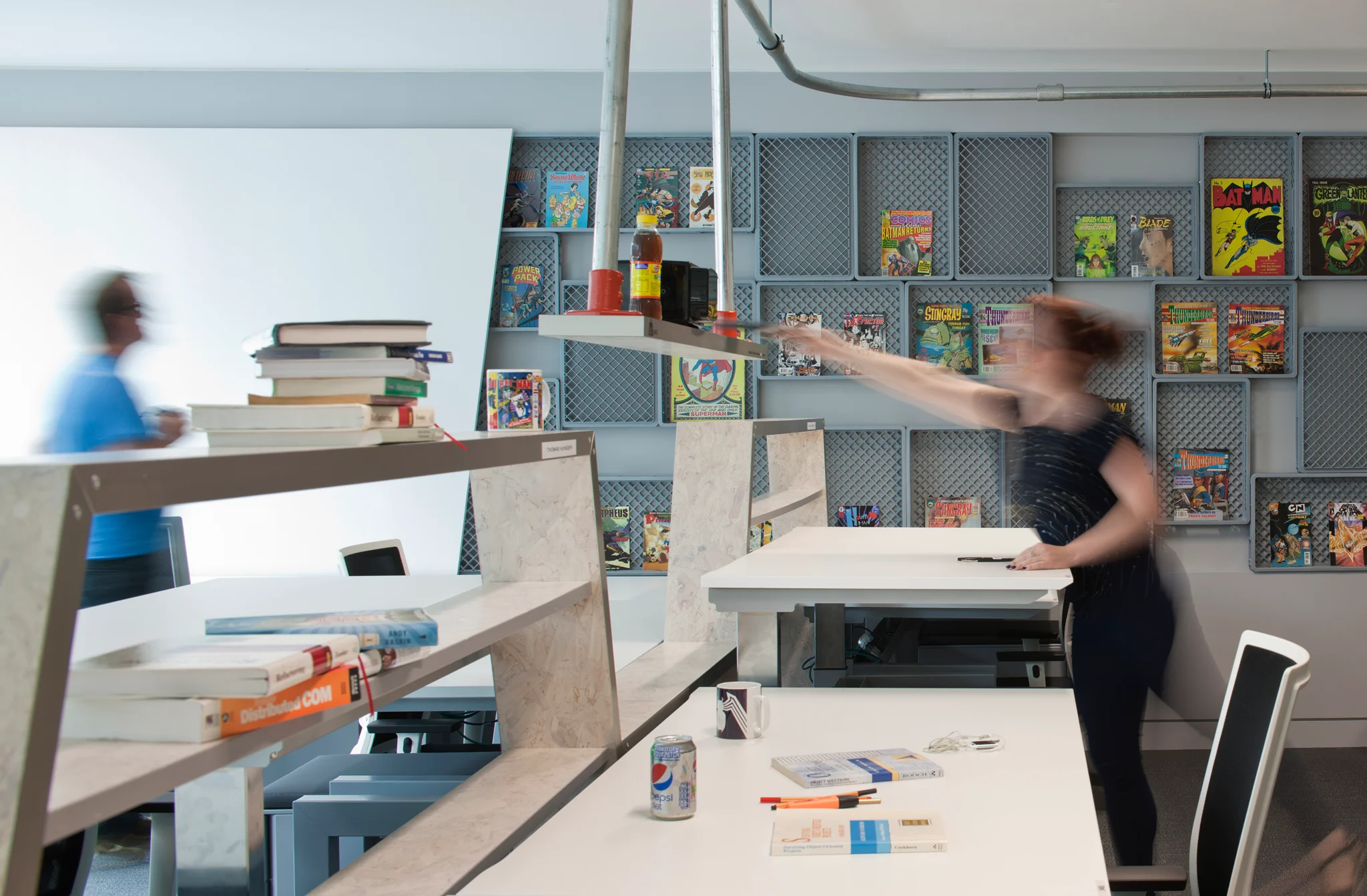 A woman using white sit/stand desks with an array of colourful books on top, complemented by wooden & industrial-style shelving with more books on