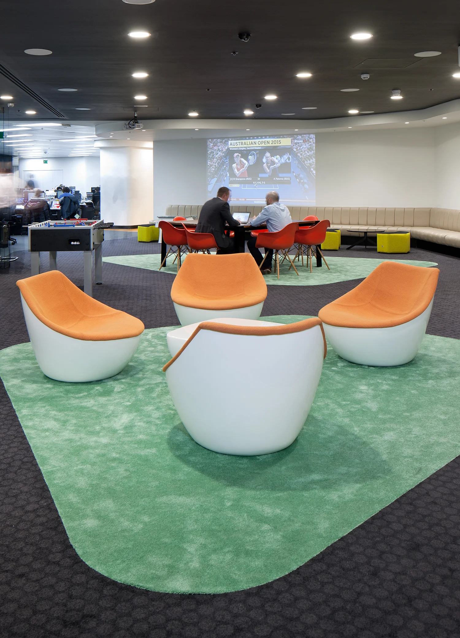 white and orange tub chairs on a green rug. Two men work in the background