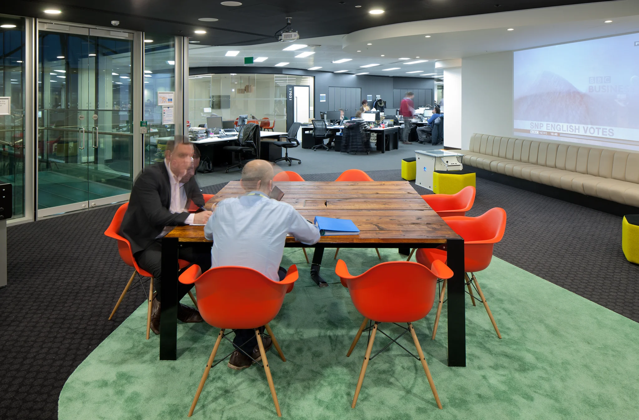 two men sat at a wooden table with red chairs on a green rug