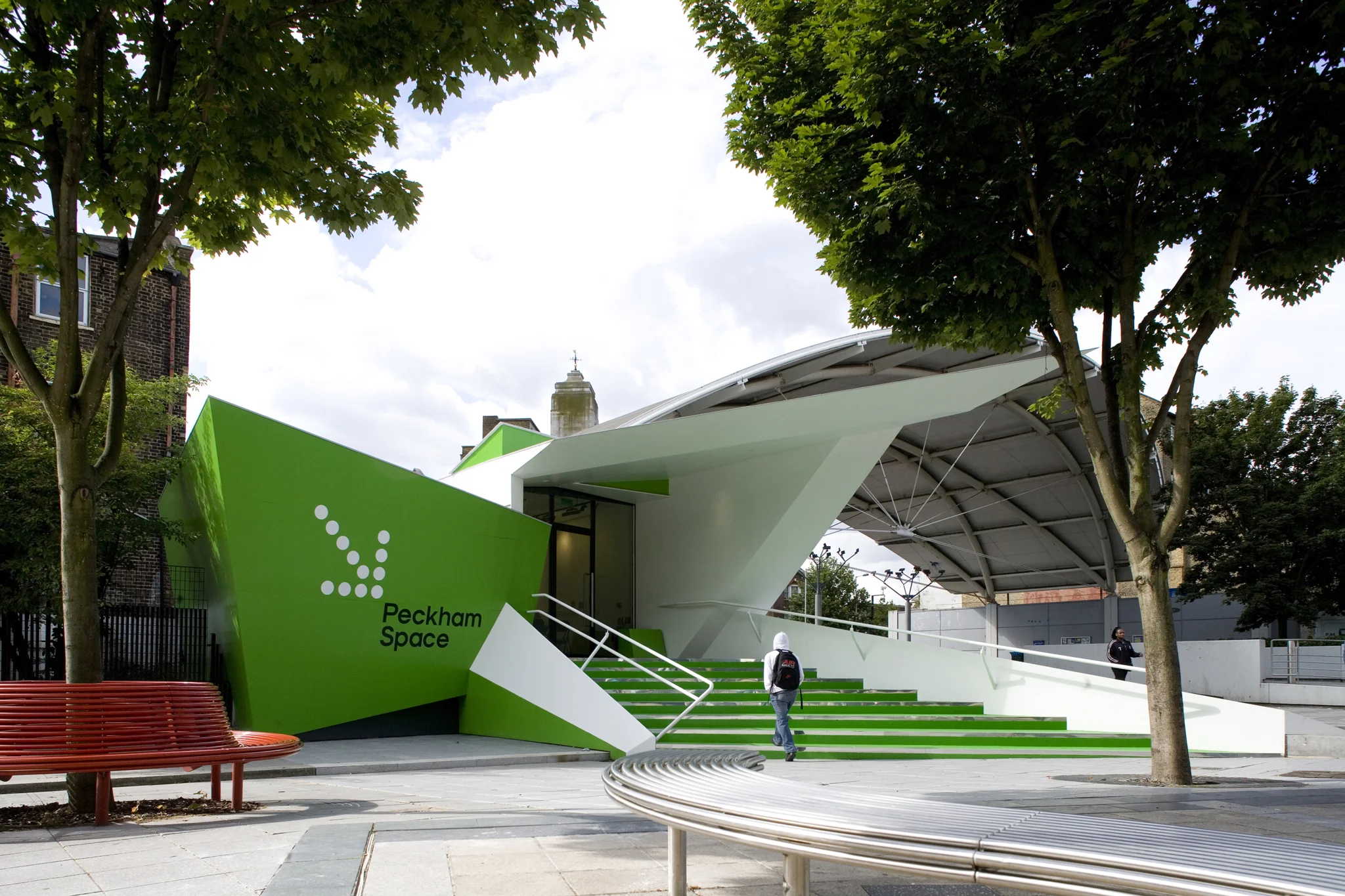 wide angled exterior shot of a diagonal view of  the green entrance to Peckham space with a white arrow pointing towards the door. Trees & the sky frame the shot