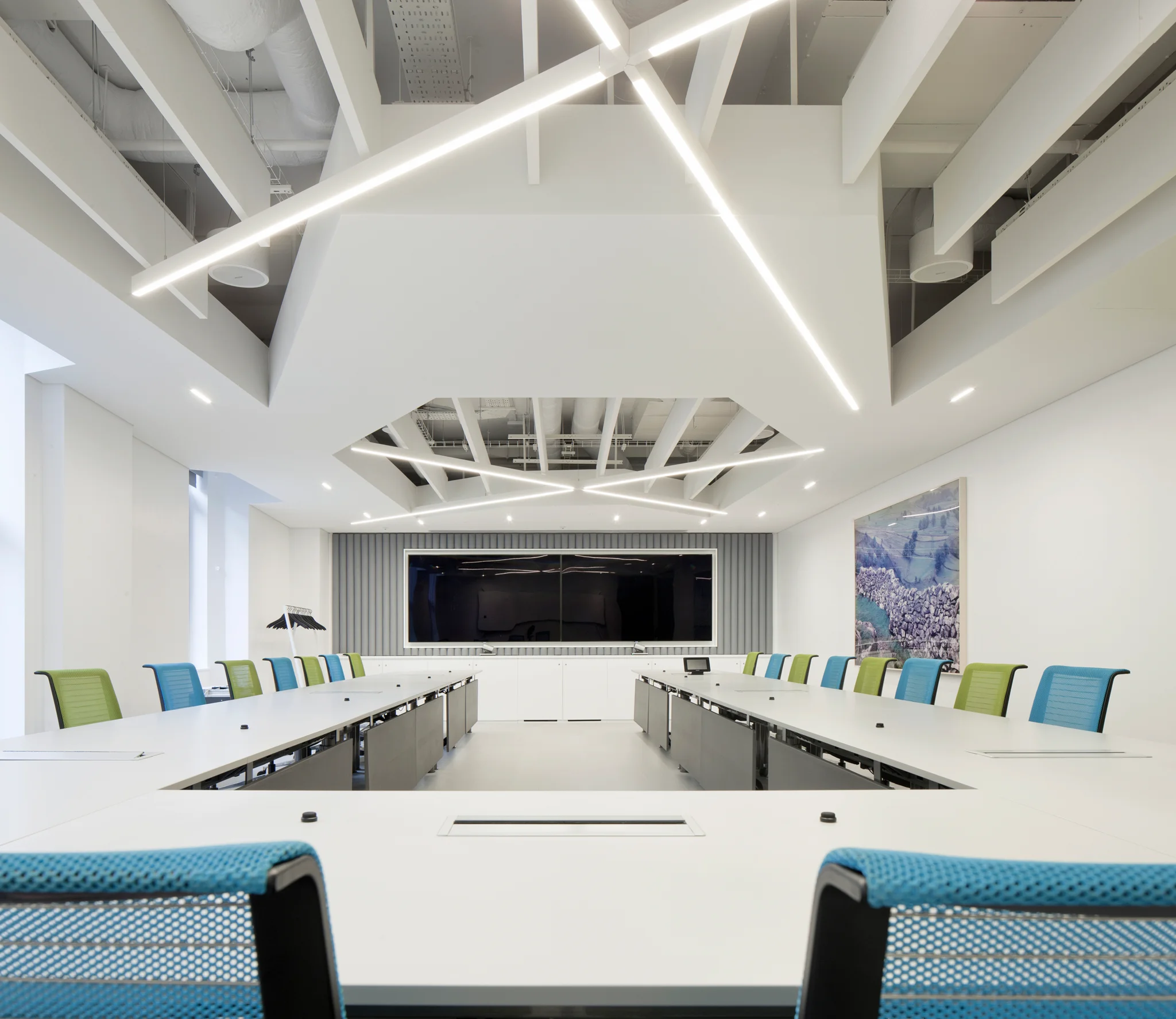 A completely white board room with white strip lighting set into a cross shape on the ceiling. Green & blue chairs flank either side of the white u-shaped table
