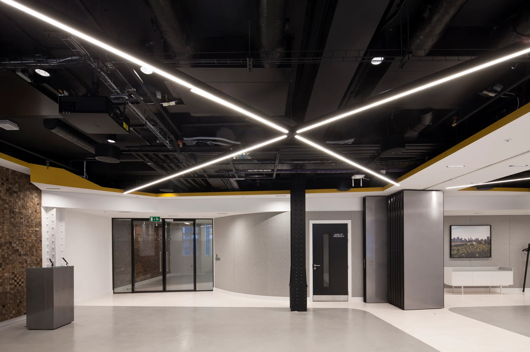 An open event space with dark exposed ceiling, white LED strip lighting in a cross shape, a lectern and movable wall revealing artwork on a wall