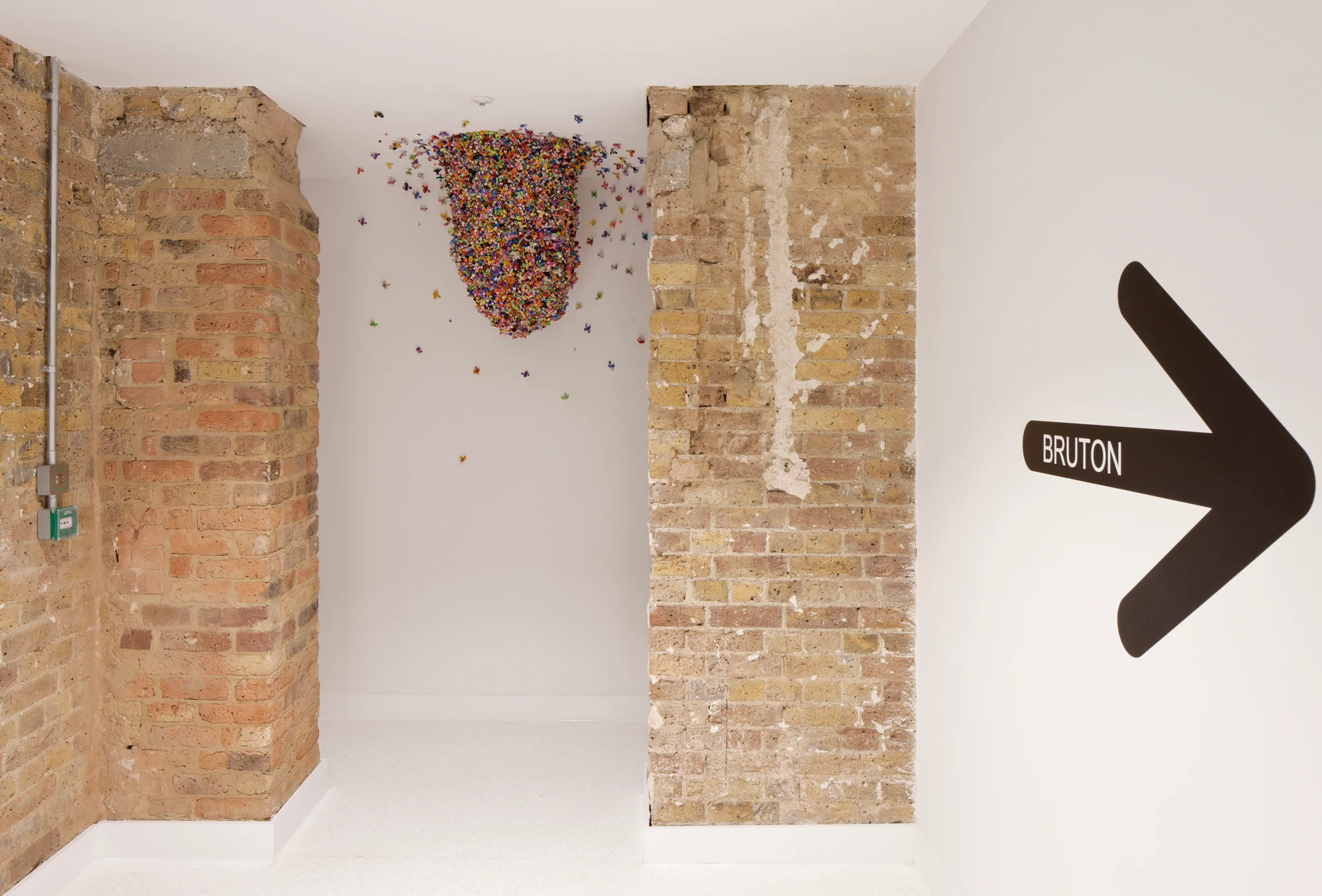 exposed brickwork protrude either side of a 3D piece of artwork of a collection of tiny multicoloured glass bees set into a 'hive' shape. A black arrow pointing to the right is set on a white wall