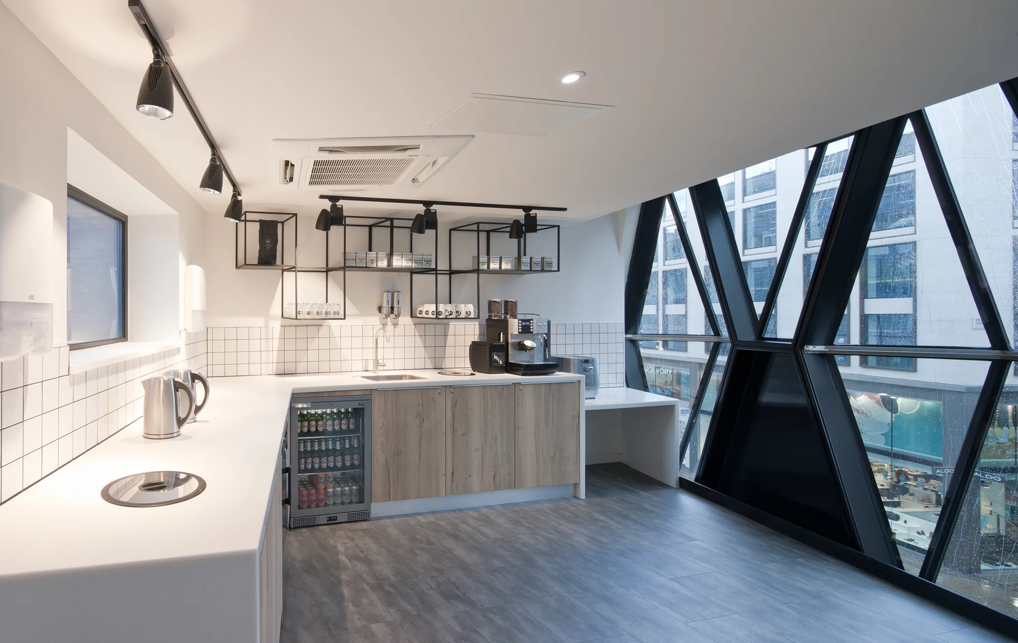 A white modern designed kitchen tea point to the left with wood floors, wood fronted cupboards and black exposed shelving adjacent to glass windows that look out on to Oxford street
