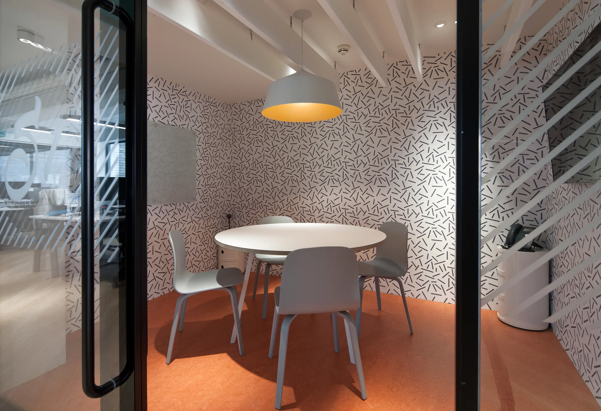 A meeting room with burnt orange coloured carpet, walls with white & black haphazard lines, a central large white drop pendant with yellow interior suspended over a white table & light blue chairs