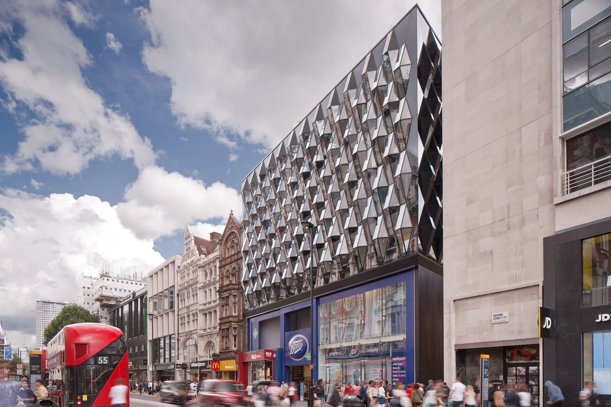 A busy street with traffic and people looking over to an exterior shot of the Co-work building from street level, showing the bevelled glass facade over Boots' shop