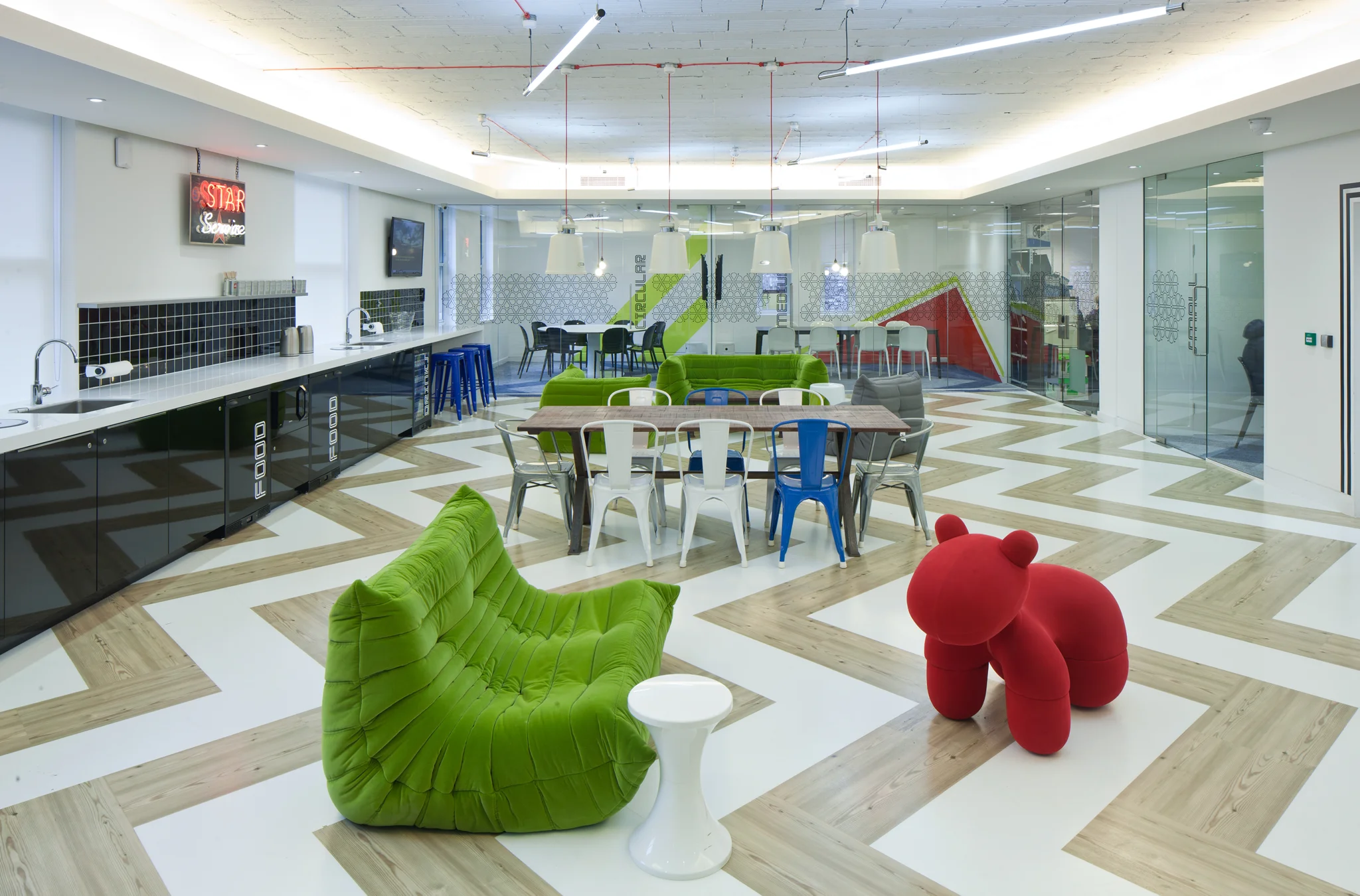 side view of kitchen area with black tiling & cupboard fronts, chevron style white and wooden with a neon sign on the wall and with two white pendant lamps suspended over furniture