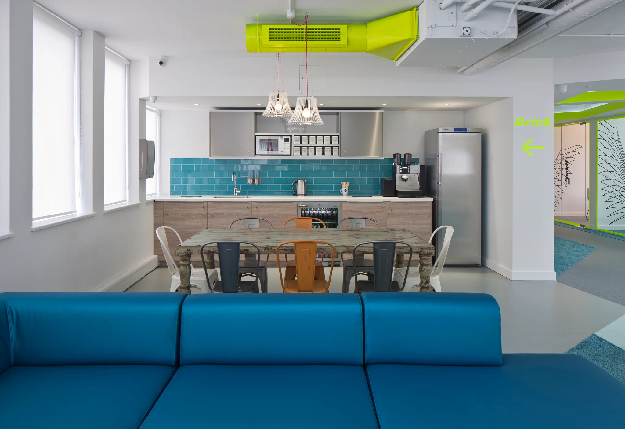 a tea point with blue sofa seating in the foreground and a table and chairs with a collection of suspended lamps and kitchen units behind