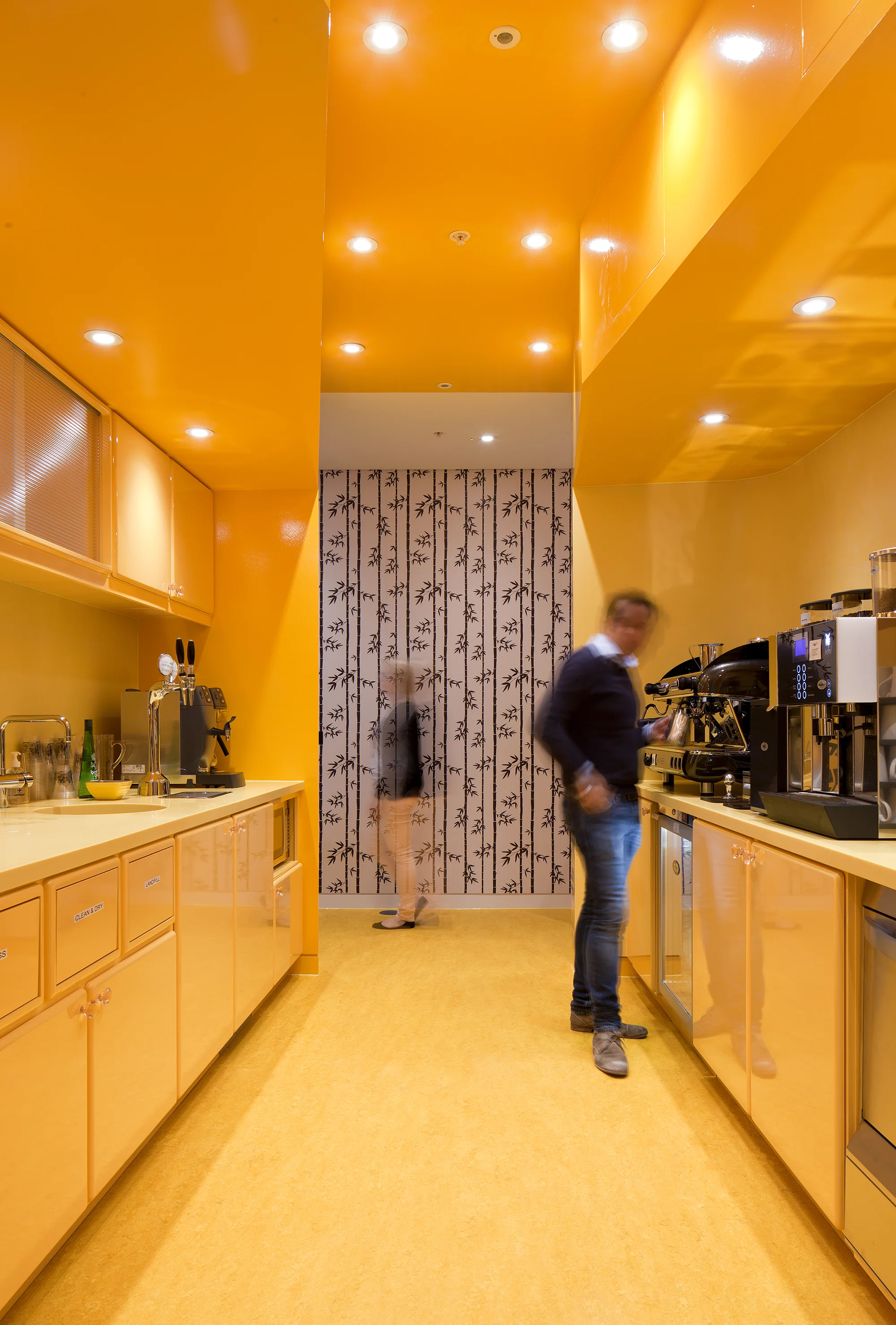 a completely yellow kitchen with a man stood by coffee machines and kitchen counter tops. a doorway opens to a black and white printed wall with a woman walking through