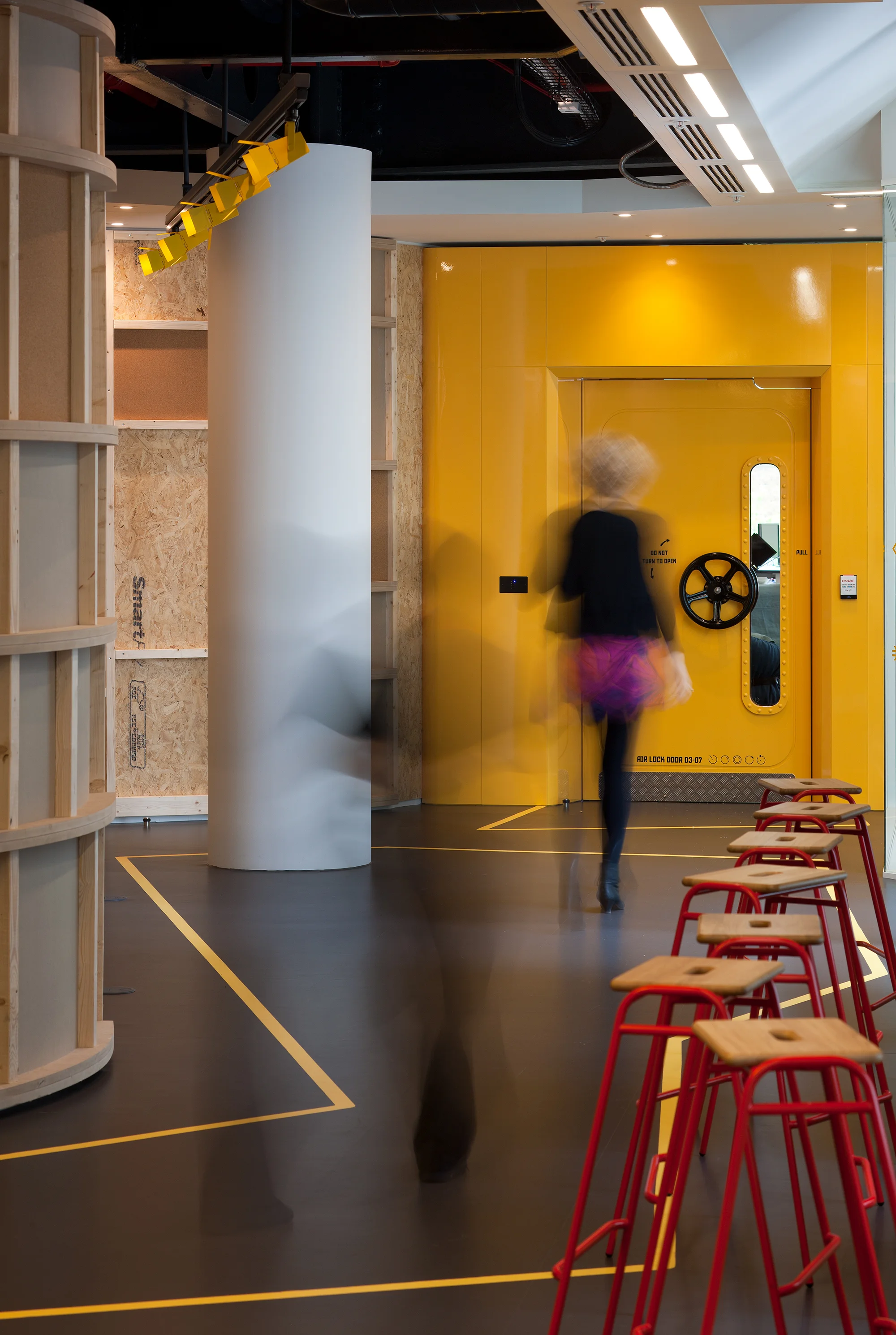 A woman walks through a corridor space with exposed stud work chipboard walls to the left and a yellow wall and doorway with a black circular wheel as the handle