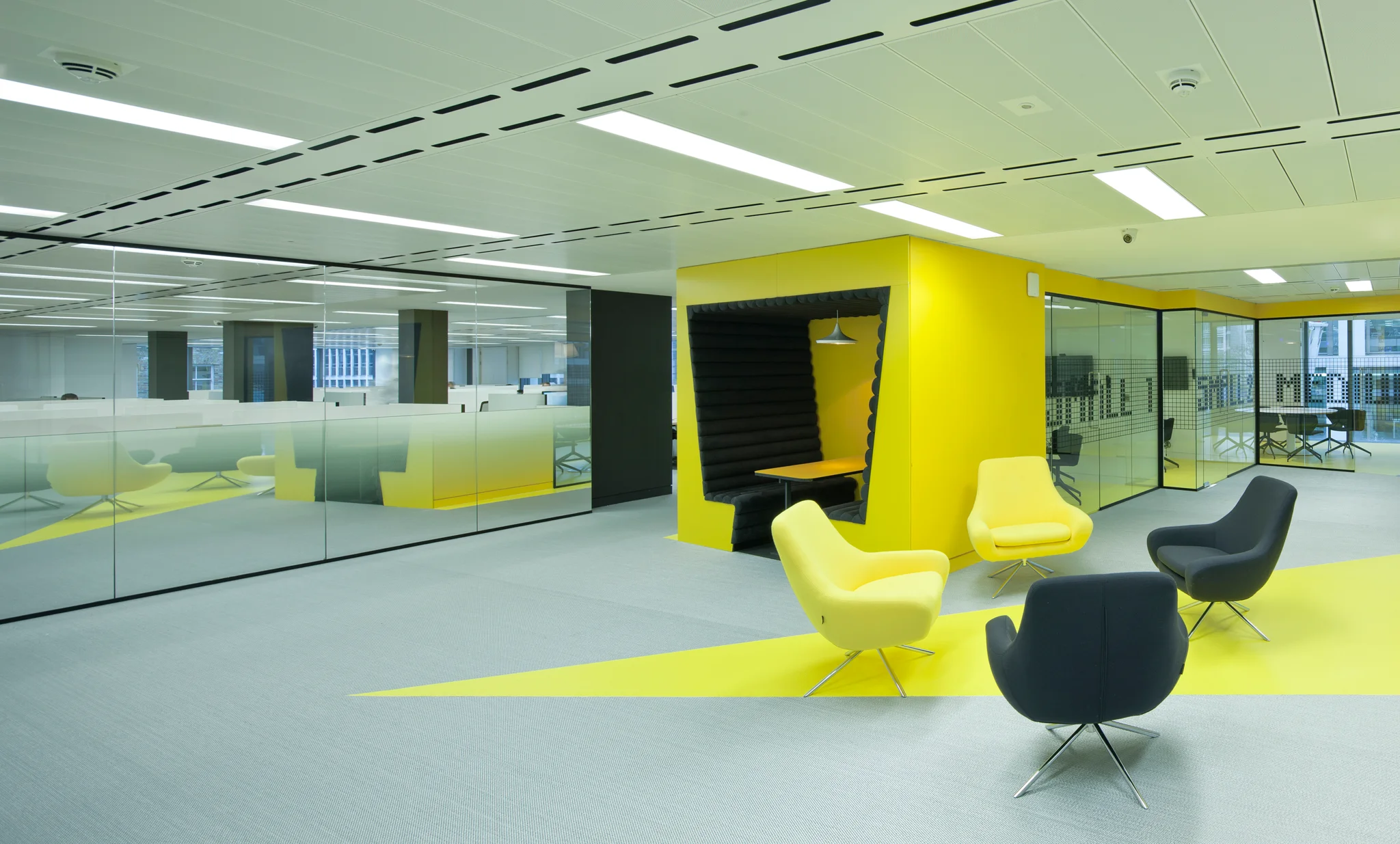 a workspace with a side view of a bright yellow booth with black seating inside. Yellow paint extends diagonally across a grey floor. Yellow and black chairs sit next to the booth