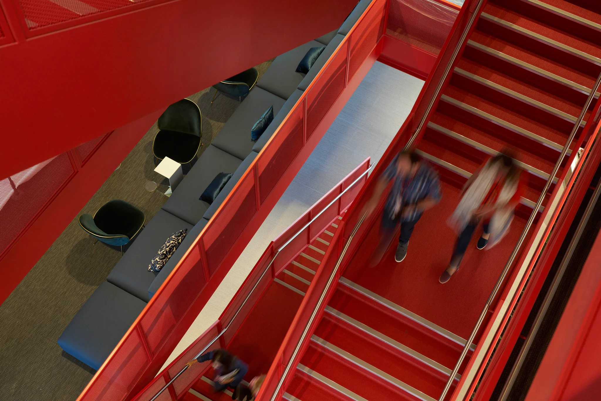 an image looking down a bright red staircase from above - showing the levels of floors. Two people walk down the stairs while the left hand side of the image shows dark blue seating areas & chairs