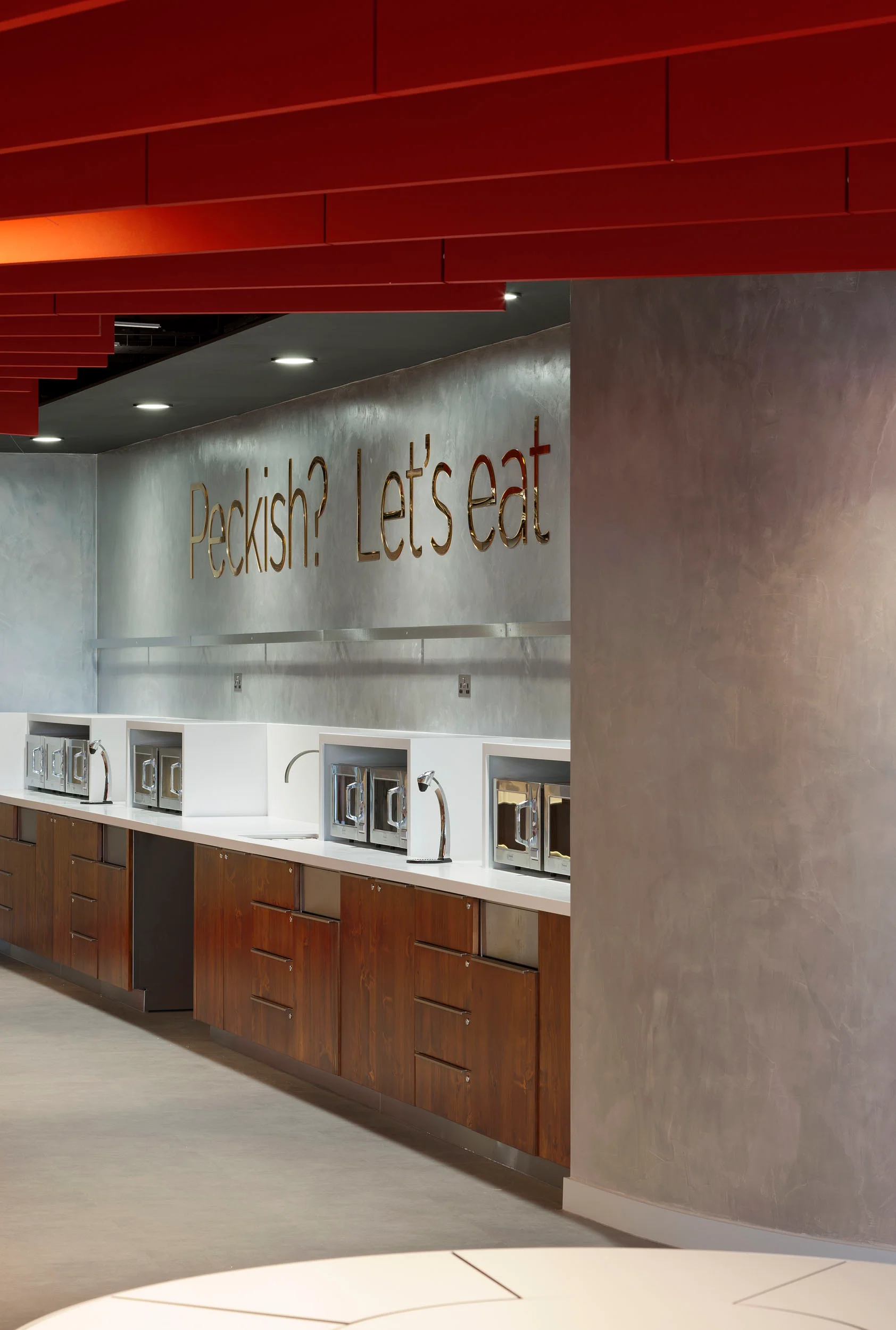 a close up shot of a kitchen tea point with multiple taps and microwaves on the counter. Gold letters on the concrete-effect wall above read: 'Peckish? Let's eat'