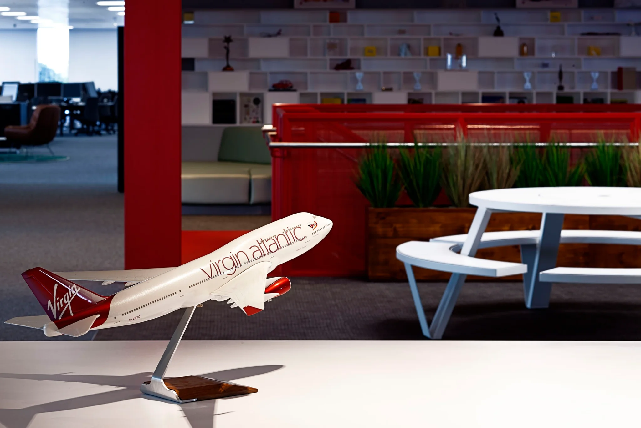 A close up shot of a model Virgin Atlantic aeroplane sat on a counter top with a circular white bench table and red staircase & planting in the background