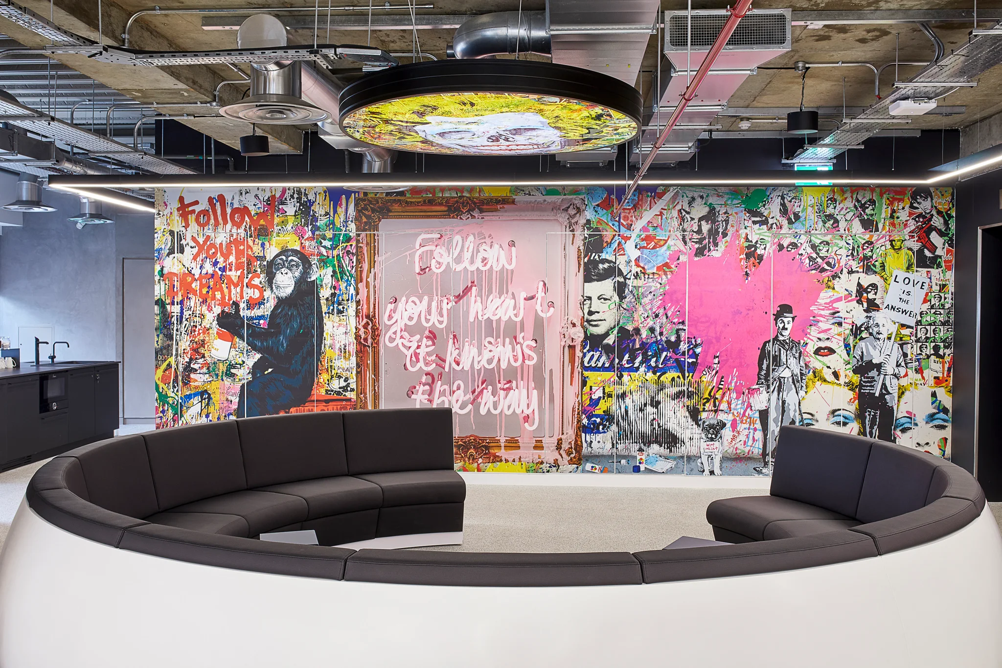 A large black and white round sofa with circular yellow artwork suspended above. A whole wall of bright pink artwork is behind