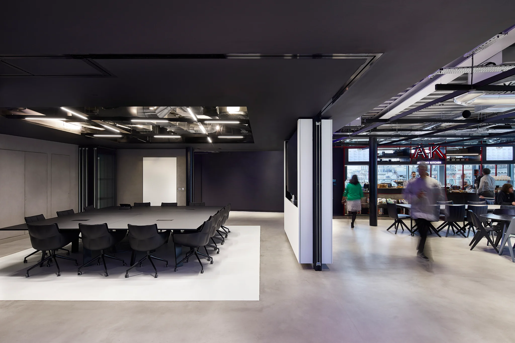 Black large table and chairs against grey and white floor/walls of a meeting room. The wall is pulled back to reveal open space. People are using the space on the right hand side