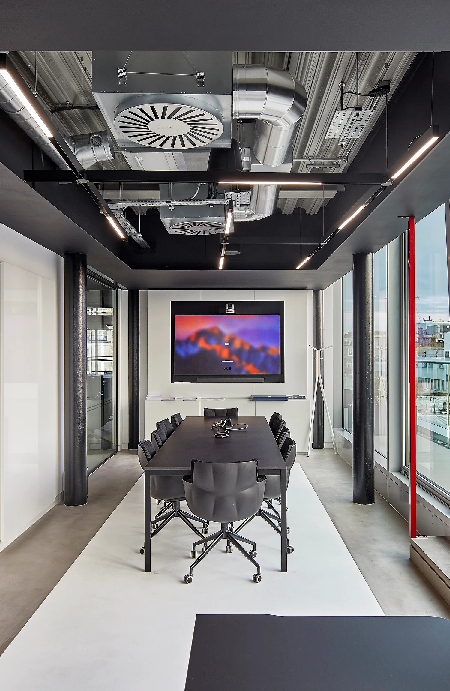 Meeting room with black table & chairs, a white floor & red structural detailing. TV screen at the end of the room