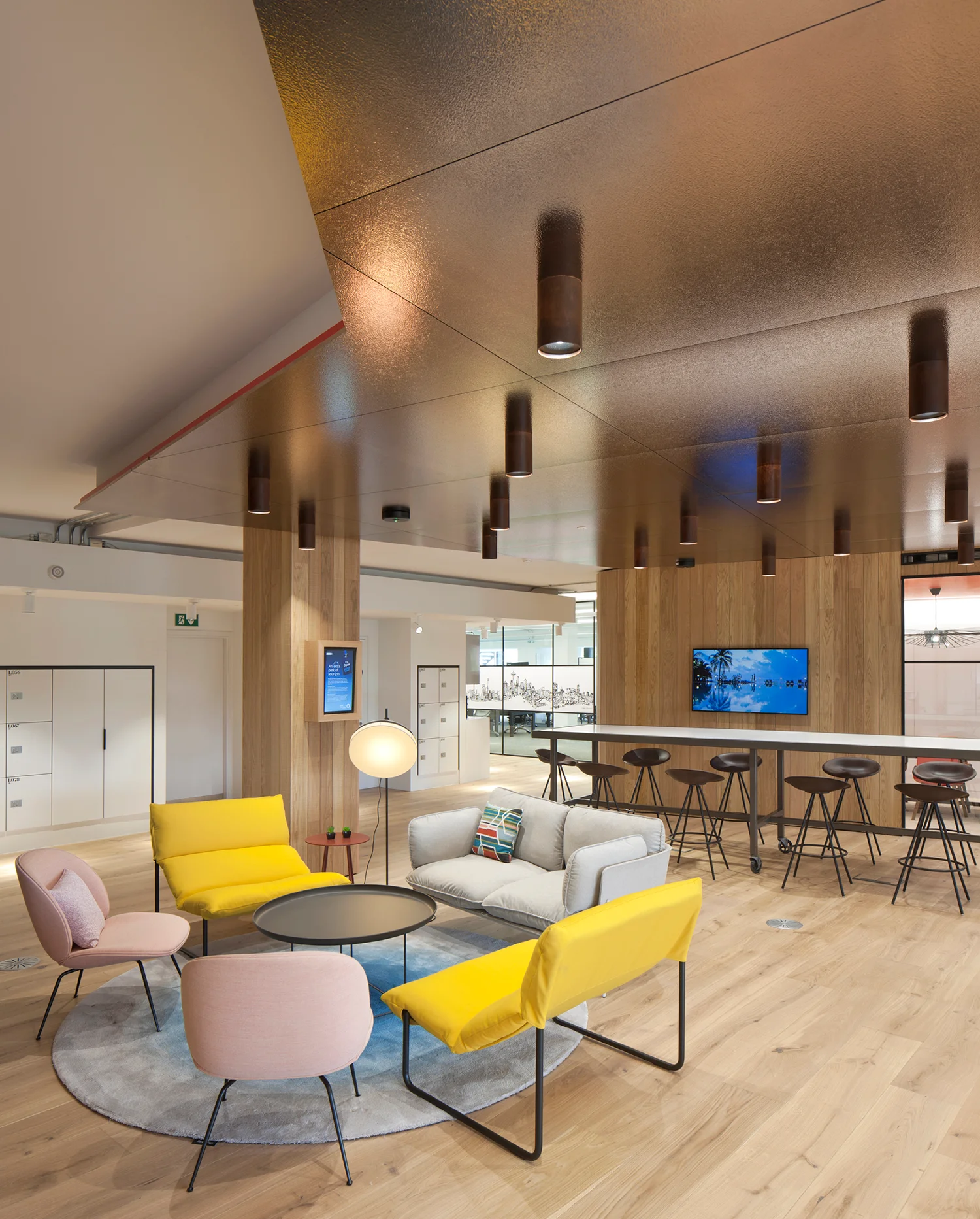 Gold ceiling, wooden floor & walls with bright yellow, light pink chairs and light grey sofa in an arrangement in front of high table and chairs