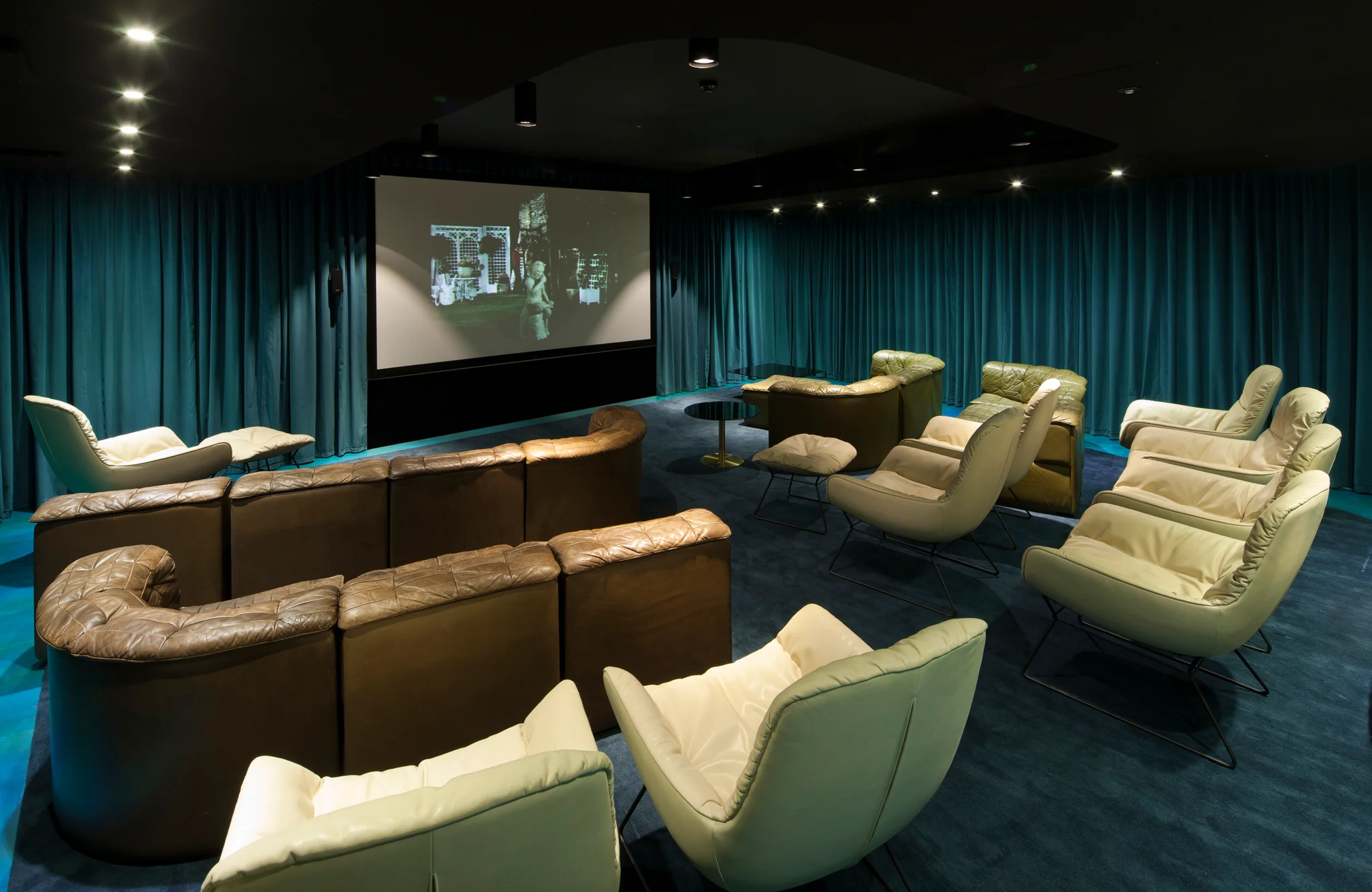 A cinema room with teal coloured walls & carpet. Beige and brown retro leather sofas are arranged facing a large cinema-style screen