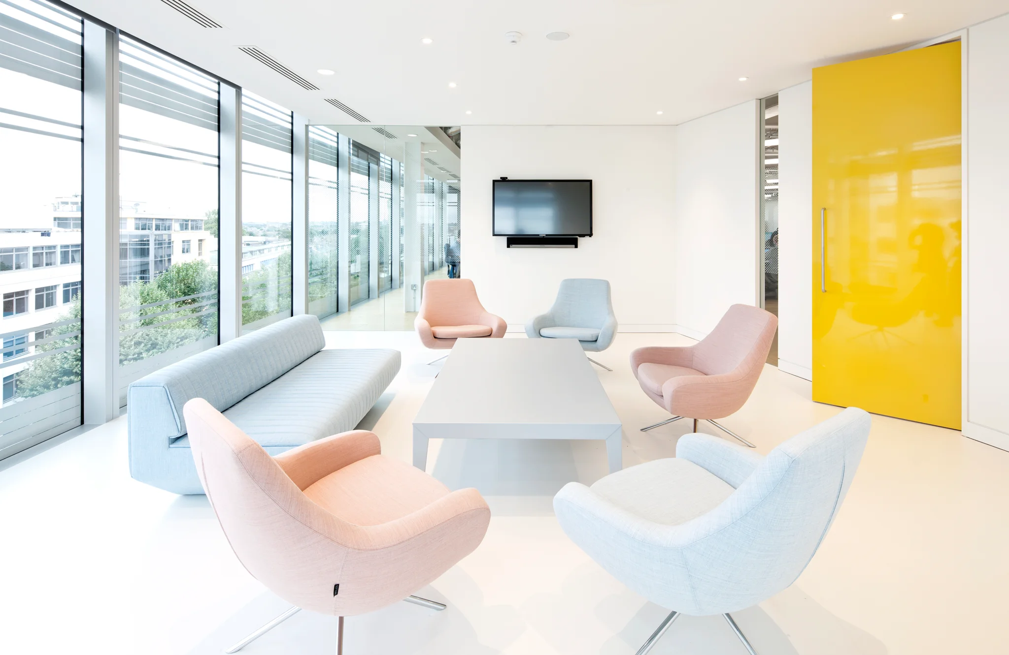 A white meeting room with a bright yellow door and light grey and pink sofas around a white table